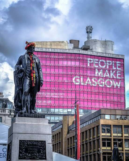Glasgow statue with tartan hat and scarf in front of a large building clad in bright pink wrap around banner proclaiming:
'People Make Glasgow.'