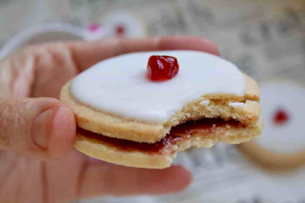 Image of an empire biscuit. White icing on top with a cherry. Biscuit has a bite taken from it, showing the red jam filling between the layers.